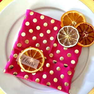 May include: A pink polka dot napkin with gold polka dots and a dried orange slice with a name tag that says "Claire" on it. There are also dried citrus slices on the napkin.