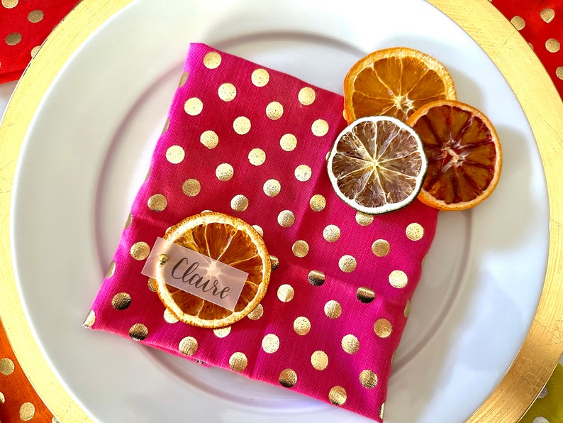 May include: A pink polka dot napkin with gold polka dots and a dried orange slice with a name tag that says "Claire" on it. There are also dried citrus slices on the napkin.