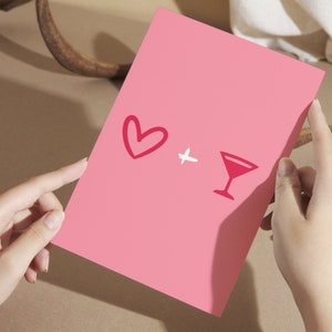 May include: A pink card with a red heart and a red cocktail glass drawn on it. The card is being held by a person's hands.