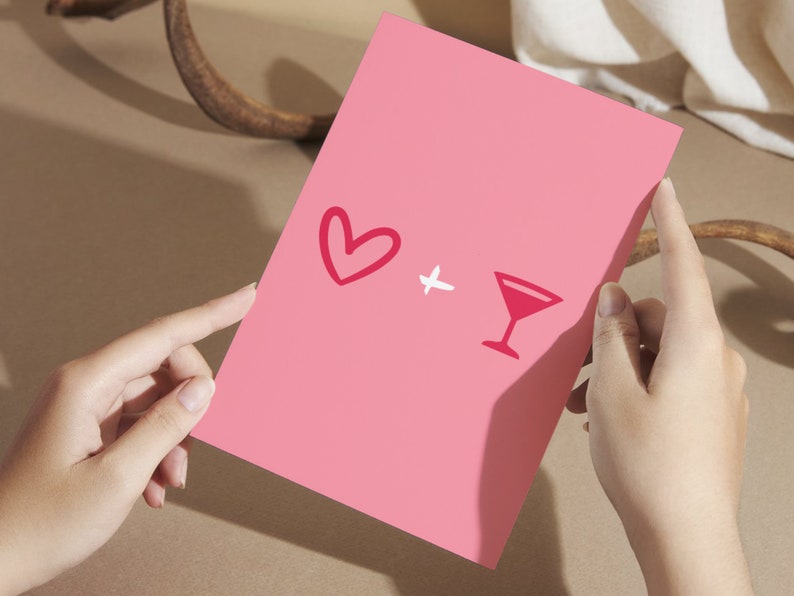 May include: A pink card with a red heart and a red cocktail glass drawn on it. The card is being held by a person's hands.