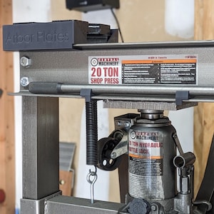 May include: A grey metal shop press with a 20-ton capacity. The press has a black handle and a black spring. The press is labelled "Central Machinery 20 Ton Shop Press". The press is sitting on a wooden surface. The image also includes a black metal plate labelled "Arbor Plates".