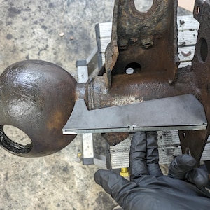 May include: A close-up of a rusty metal bracket with a metal plate attached to it. The plate is being held by a gloved hand.