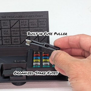 May include: A black fuse box with a clear lid. The lid has a fuse puller built in and a chart showing the fuse amperage for each circuit. The box also has a compartment for spare fuses.