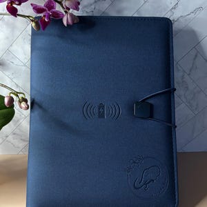 Op de afbeelding: Een marineblauw notitieboek met een draadloos oplaadsymbool en een olifantenlogo. Het notitieboek heeft een bandsluiting en is versierd met een paarse orchidee. Het notitieboek is ontworpen voor dagelijks gebruik.