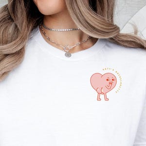 Op de afbeelding: Wit T-shirt met een roze hartgraphic met benen en een knipoog. Het hart heeft de tekst "Make it a great day" erboven.