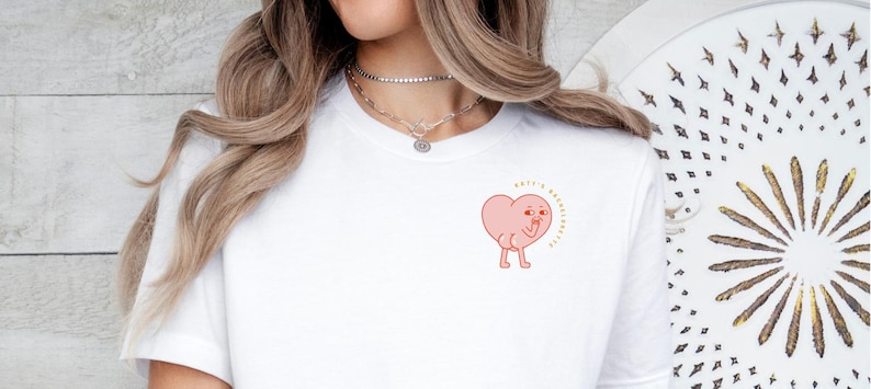 Op de afbeelding: Wit T-shirt met een roze hartgraphic met benen en een knipoog. Het hart heeft de tekst "Make it a great day" erboven.