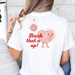 Op de afbeelding: Wit T-shirt met een rode hartgrafiek en de tekst "Bach that a up!" en "Katy's Bachelorette".