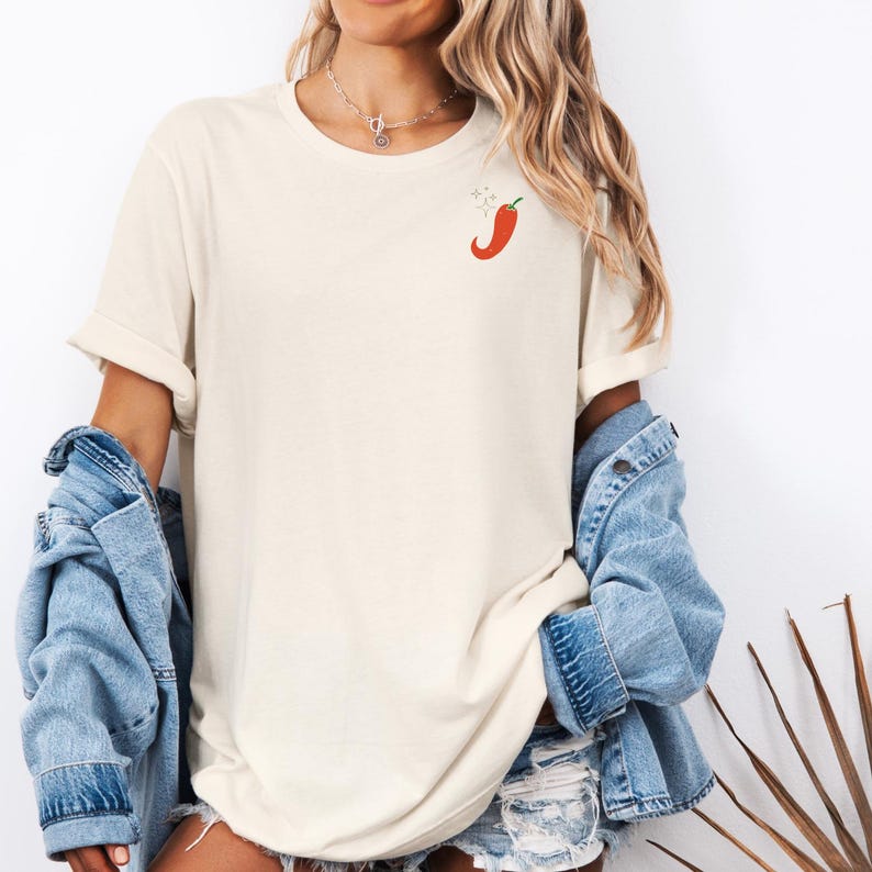 May include: A white oversized t-shirt with a red chili pepper graphic on the left chest. The model is wearing a denim jacket and denim shorts.