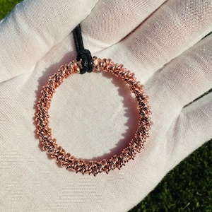 May include: A copper-colored pendant necklace with a circular design. The pendant is made of tightly coiled wire, creating a textured surface. It hangs from a black cord. The necklace is displayed against a white background.