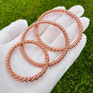 May include: Three copper wire bracelets with a twisted design. The bracelets are stacked on top of each other and are displayed on a white surface.