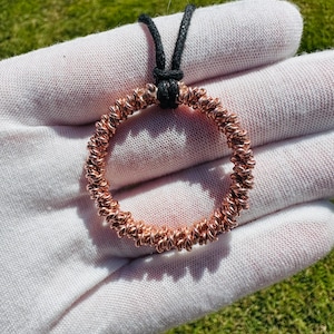 May include: A copper-colored pendant necklace featuring a circular design made of tightly coiled wire. The pendant hangs from a black cord. The necklace is displayed against a white background with green grass visible.