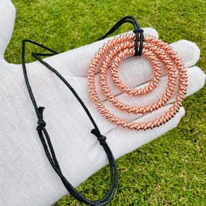 May include: A copper-colored pendant necklace featuring three concentric spiral rings. The rings are suspended from a black cord. The necklace is displayed against a green grassy background.