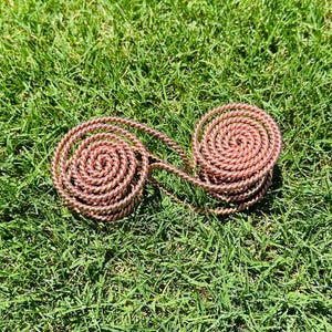 May include: Two copper-coloured spiral coils connected by a twisted wire, resting on green grass. The coils are tightly wound, creating a circular shape. The design is symmetrical and visually appealing.