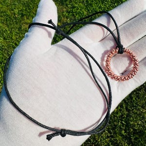 May include: A rose gold colored wire pendant necklace with a circular design. The necklace is on a black cord.