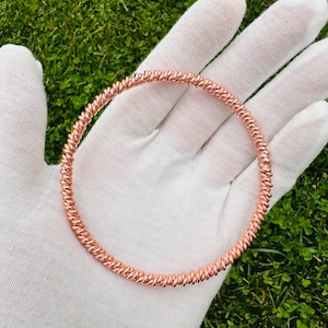 May include: A copper-colored, spiral-textured bracelet. The bracelet is circular and appears to be made of twisted metal. The bracelet is held in a gloved hand against a background of green grass.
