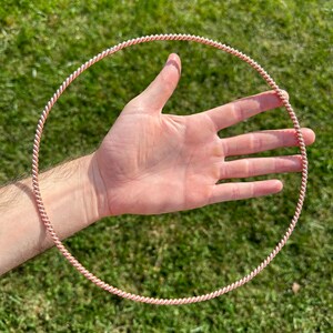 May include: A copper-colored, twisted metal ring with a pink accent held in a hand against a green grass background. The ring is circular and appears to be made of wire. The twisted design gives it a textured appearance.