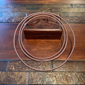 May include: Three copper-colored, twisted metal hoops rest on a wooden surface, partially obscuring a small, dark wooden box with an engraved design. The hoops are arranged in a nested fashion, creating a layered effect.