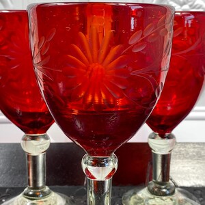 May include: Three ruby red glass goblets featuring a clear glass stem and base. Each bowl is decorated with an etched floral pattern consisting of a central daisy-like flower surrounded by delicate leafy vines and small circular accents.