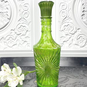 May include: A vibrant green glass decanter featuring a sunburst pattern on the body and a matching fluted stopper. The bottle has a slender neck and a wide base, with a textured surface that catches the light. A small white flower sits nearby.