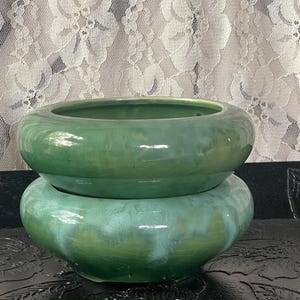 May include: Two stacked, round, green ceramic planters with a glossy finish. The top planter is a lighter shade of green than the bottom one. The planters are sitting on a black surface.