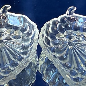 Könnte beinhalten: Zwei klare Glasservierschalen in Form von Trauben. Jede Schale hat eine strukturierte Oberfläche und einen stielartigen Griff. Die Schalen stehen vor einem dunkelblauen Hintergrund, der das Licht reflektiert und einen funkelnden Effekt erzeugt.