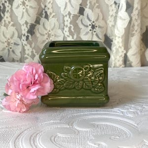 May include: A green ceramic square vase with a floral relief design. A pink carnation flower rests beside the vase. The vase has a glossy finish and a rectangular opening at the top.