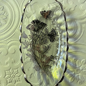 Könnte beinhalten: Ovale Glasschale mit silbernem Rand und floralem Design. Die Blumen sind schwarz und silber, mit braunen Akzenten. Die Schale hat eine strukturierte Oberfläche und dient wahrscheinlich dekorativen Zwecken.