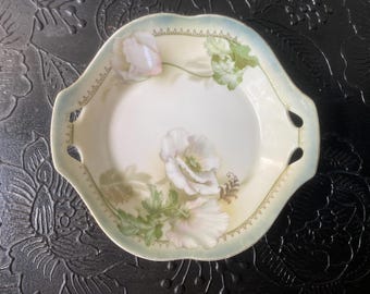 May include: An antique, hand-painted porcelain serving dish. The dish features a white interior with a floral design of white and pink poppies and green leaves. The rim is a pale yellow with a light blue border and decorative cut-out handles.