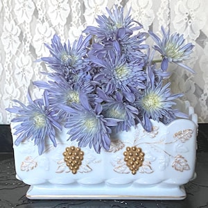 Puede incluir: Un jarrón rectangular de cerámica blanca con detalles dorados, con un diseño de uvas y lleno de flores azules. El jarrón tiene un borde festoneado y está colocado sobre una superficie oscura.