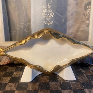 May include: A gold and white ceramic dish shaped like a fish. The dish has a white interior and a gold exterior with a textured, wavy design. The dish is sitting on a white stand.