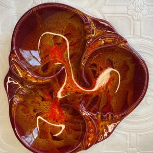 Puede incluir: Un plato de cerámica vintage de tres secciones con una forma orgánica única. El plato presenta un esmalte brillante en tonos burdeos, naranja y amarillo, con detalles en blanco. El diseño recuerda a flujos de lava o arte abstracto.