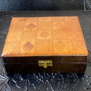 May include: A square, vintage-style box with a brown leather exterior. The lid features a repeating pattern of embossed geometric shapes in a lighter shade of brown. The box has a dark brown base and a brass clasp.