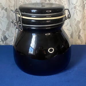 May include: A black ceramic jar with a rounded body and a matching lid. The lid is secured with a metal clasp and a white rubber seal. The jar has a glossy finish and reflects the surrounding environment.