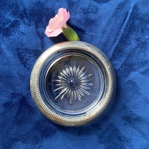 Peut inclure: Un dessous de verre rond en verre avec un bord argenté et un motif en étoile. Une œillet rose repose sur le dessous de verre, sur fond bleu. Le bord du dessous de verre est décoratif.