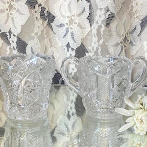 Peut inclure: Deux sucriers en verre taillé transparents, ornés d'étoiles et de motifs géométriques. L'un est muni de poignées. Ils reposent sur une surface réfléchissante, avec des fleurs blanches et des rideaux en dentelle en arrière-plan.