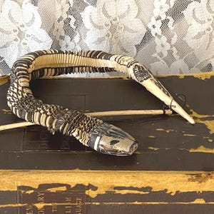 May include: A handcrafted, articulated snake figurine with a detailed black and cream pattern. The snake is posed on a stack of antique books, with a lace backdrop. The snake's head is painted with black and white details.