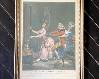 Könnte beinhalten: Ein gerahmter antiker Druck mit dem Titel "Tight Lacing, or Fashion before Ease". Der Druck zeigt eine Frau in einem rosa Kleid, die von einem Mann geschnürt wird, mit einer weiteren Frau und einem kleinen Hund. Der Rahmen ist goldfarben.