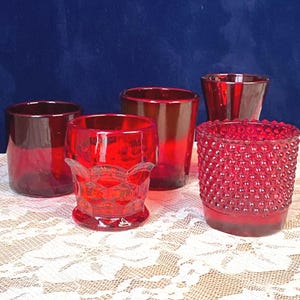 Pode incluir: Uma coleção de cinco copos de vidro vermelho. Os copos variam em forma e textura, incluindo superfícies lisas, estampadas e texturizadas. Estão dispostos sobre uma toalha de renda branca, contra um fundo azul escuro.