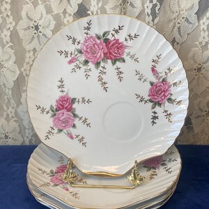 May include: A set of white, shell-shaped plates with a gold trim and pink rose floral pattern. The plates are stacked on a gold-coloured metal stand. The background is a lace curtain.