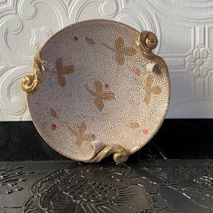 Puede incluir: Un plato decorativo y redondo con una superficie texturizada de color blanquecino. Diseños y detalles de mariposas doradas adornan el plato, junto con pequeños detalles rojos y turquesas. El plato tiene asas doradas en forma de voluta.