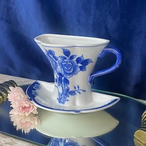 Puede incluir: Juego de taza y platillo de té de porcelana blanca y azul. La taza presenta un diseño floral azul y un asa azul. El platillo ovalado complementa la taza. El juego se exhibe sobre una superficie espejada.
