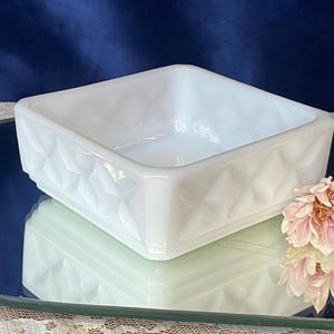 Puede incluir: Un plato cuadrado de vidrio blanco con un patrón geométrico de diamantes en los lados. El plato está sobre una superficie reflectante, con un fondo borroso y flores rosas.