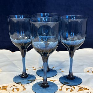 May include: A set of four translucent blue glass wine glasses with smooth, rounded bowls, slender stems, and circular bases. The glassware features a consistent deep blue hue throughout, with a clear, reflective surface and a simple, elegant design.