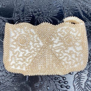 May include: A rectangular clutch bag covered in cream-coloured beads arranged in intricate floral and geometric patterns. The bag features a scalloped top edge and a small, matching beaded handle on the right side, creating a textured surface.