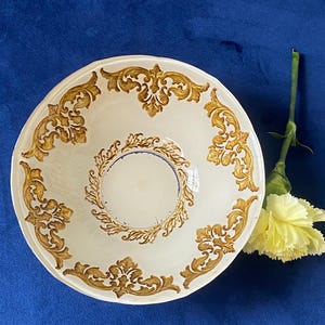 May include: A white ceramic bowl with a gold ornate design around the rim and center. A yellow flower and stem are placed to the side of the bowl against a blue background. The bowl has a circular shape.