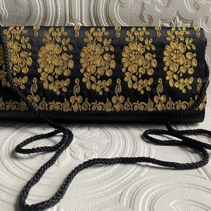 May include: Black clutch bag with gold floral embroidery. The rectangular bag has a braided black strap. The gold embroidery is in a floral pattern across the front. The bag is closed with a flap.