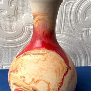 Puede incluir: Un jarrón de cerámica hecho a mano con un diseño veteado único. El jarrón presenta una base de color crema con patrones arremolinados de naranja, rojo y gris. La parte superior y el cuello son de color crema sólido, y el jarrón tiene un cuerpo redondeado y un cuello estrecho.