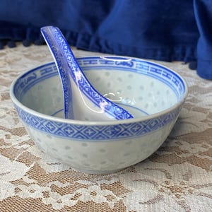 Puede incluir: Un juego de cuenco y cuchara de porcelana blanca y azul. El cuenco tiene un delicado patrón de puntos y un borde geométrico azul. La cuchara descansa dentro del cuenco, con un diseño azul a juego. El juego está sobre un mantel de encaje.