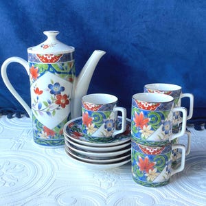May include: A white ceramic tea set with a floral design. The set includes a tall teapot, several teacups, and saucers. The cups and teapot are decorated with red, blue, and green floral patterns.