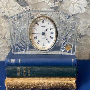 Puede incluir: Un reloj de cuarzo de cristal transparente con un bisel dorado y números romanos, marcado "Stinger". El reloj está enmarcado en un marco de cristal decorativo con diseños de hojas. Descansa sobre una pila de tres libros con cubiertas azules, verdes y doradas.